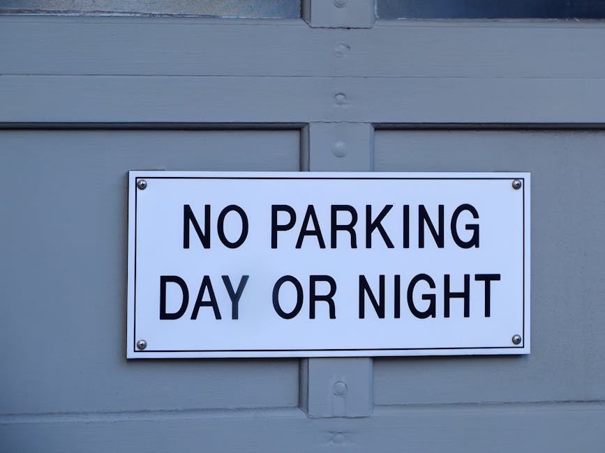 Close-up view of a white rectangular sign with bold black lettering that reads 'NO PARKING DAY OR NIGHT', mounted on a grey wooden door or panel. The sign is secured with four bolts, one at each corner. The surrounding surface appears to be painted grey wood, likely part of a house or building exterior. The image captures the sign in natural lighting, emphasizing its clear message, which is relevant for house removals and moving logistics to avoid parking fines during relocation or furniture transport activities, as managed by Man with Van North Sheen.