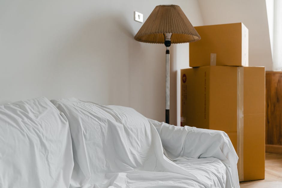 Inside a residential room prepared for a home relocation, there is a white fabric-covered sofa positioned along a light-colored wall, with visible wrinkles on the upholstery. Next to the sofa, three cardboard boxes of varying sizes are stacked, with the largest at the bottom and the smaller ones on top, sealed with brown packing tape. Behind the boxes, a tall floor lamp with a brown pleated fabric lampshade and a metallic stand stands adjacent to a wooden cabinet or side table. The environment is well-lit with natural light coming from a window out of view, highlighting the generally tidy interior ready for furniture transport or packing and moving activities. Man with Van North Sheen’s services are implied through the scene of furniture and box preparation for a house move in North Sheen, SW14, during a typical loading process at a residential property.