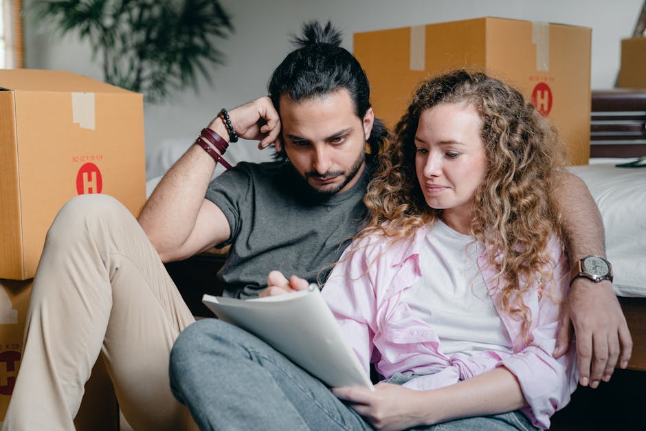A man with long dark hair tied in a bun and a beard, wearing a dark grey t-shirt and beige trousers, sits on the floor inside a room with moving boxes, carefully examining a clipboard or list related to house removals. Next to him, a woman with curly blonde hair, dressed in a light pink shirt, also sits on the floor and looks at the same document. They are surrounded by packed cardboard boxes sealed with packing tape, some of which bear red labels and printed branding, indicating they are prepared for a home relocation. Behind them, a partially visible bed and additional boxes suggest an organized packing and moving process in a residential setting. The scene depicts attention to detail during a furniture transport or home packing session, supported by various packing materials such as plastic wraps and boxes, highlighting the logistics involved in a relocation service, as offered by Man with Van North Sheen.