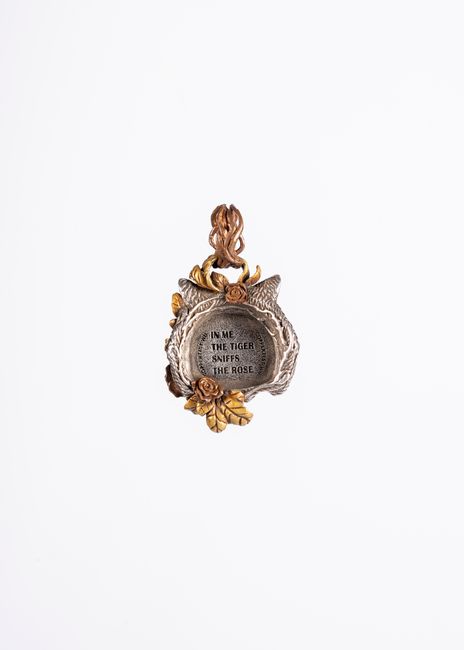 A decorative silver pendant with intricate floral details and a central circular inscription reading 'In me the tiger sniffs the rose' hangs against a plain white background. The pendant features a sm