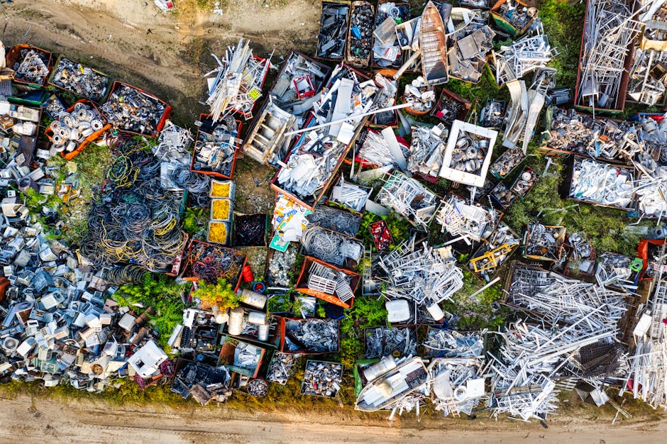 An aerial view of multiple large outdoor containers and pallets filled with scrap metal, discarded appliances, used electronic equipment, and various household items, all arranged in a scrapyard environment on a dirt surface. Some of the containers are open, revealing tangled wires, metal sheets, and broken fittings, while others are closed or partially covered with tarps. Vegetation and patches of grass are visible between the piles, and a gravel or dirt road runs along the bottom edge of the image. This scene depicts a busy scrap collection or recycling site, relevant to services like furniture transport, packing, or moving logistics offered by Man with Van North Sheen, especially in relation to clearing bulky waste during home relocation or moving projects.