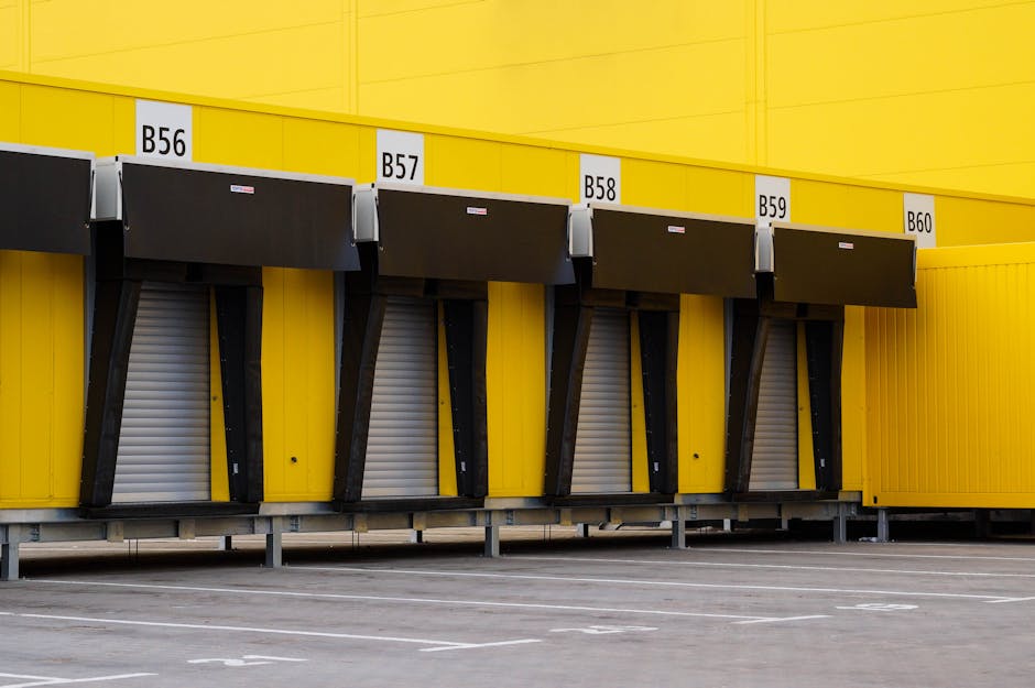 A row of four loading bays at a warehouse or storage facility, with each bay marked by white labels numbered B56, B57, B58, and B59. The bays are fitted with black retractable dock shelters to facilitate the loading and unloading process, and are positioned beneath a yellow wall. The ground in front of the bays is a paved parking area, with white lines demarcating parking spaces, and no vehicles are visible in the immediate vicinity. This environment suggests a setting for home relocation or furniture transport operations, with the bays likely used for moving goods in or out using company vehicles. The lighting is neutral, indicating daytime conditions, and the image captures the precise area where loading and unloading activities take place, relevant for moving companies such as Man with Van North Sheen that assist with packing, loading, and transportation services.