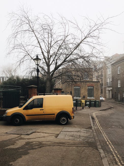 A yellow commercial van parked on a residential street near a leafless tree and a vintage-style lamppost during daytime. Behind the van, there is a brick building with barred windows and a black gate, alongside a group of four green wheelie bins placed on the pavement. To the right, a narrow road with double yellow lines curves uphill past additional residential buildings. The van is positioned close to the curb, with ample space for loading or unloading goods typically associated with home relocation or furniture transport. The setting appears calm and suitable for a moving service, with the van ready for loading furniture, boxes, or household items, consistent with house removal activities. The scene demonstrates the ease of van access and parking in a suburban environment near Kew Gardens, aligning with professional removals services offered by [COMPANY_NAME], such as packing, loading, and transport within local areas like North Sheen.