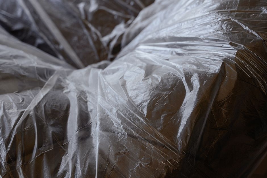 Close-up view of several large cardboard boxes wrapped in clear plastic film, stacked on top of each other inside a property, likely during a house removal or packing process. The boxes are secured with plastic wrap to protect contents during transport. In the background, part of a doorway or entrance is visible, suggesting the location may be the interior of a home preparing for a move. The lighting is natural, illuminating the packing materials and the boxes, which are situated on a flat surface, possibly a floor or a loading area. The scene reflects typical packing activities involved in home relocation, with a focus on careful preparation and secure packaging of household items. Man with Van North Sheen provides such removals services, including packing and furniture transport, as part of their house moving process, underscoring the importance of meticulous packing during a move.