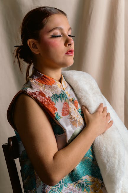 The image shows a young woman with light skin and brown hair tied back, sitting indoors with closed eyes and a serene expression. She is wearing a colorful sleeveless top with floral patterns and an orange collar, and is holding a white, fluffy object close to her chest with both hands. Behind her, beige curtains serve as a backdrop, and soft, natural lighting illuminates her face and upper body. The scene appears calm and introspective, with a subtle focus on comfort and tranquility, related to the concept of home and personal space. This image may be used in content discussing packing, moving, or personal belongings during a house relocation, with reference to furniture or household items, aligning with services like removals or home relocation, as offered by Man with Van North Sheen.