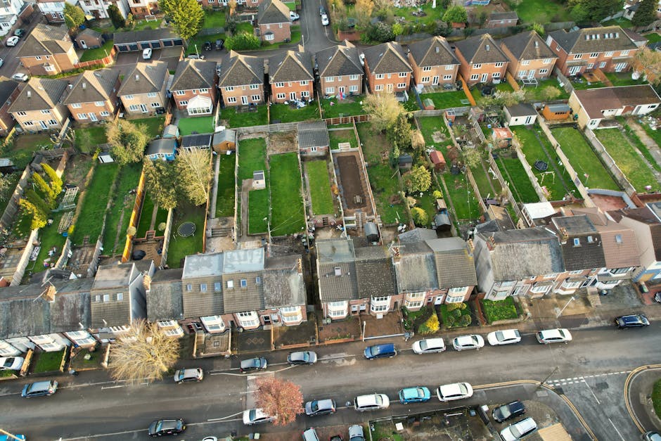 An aerial view of a residential street with a row of terraced houses at the front, each with small front gardens and parked cars along the pavement. Behind the houses, there are multiple fenced garden plots with various features such as lawns, trees, sheds, and patios. Some gardens contain outdoor furniture, play equipment, or garden tools, while others are empty or have construction materials. The scene shows a clear day with natural lighting, highlighting the general environment for house removals or home relocation services. The image illustrates the typical urban setting for furniture transport and packing and moving activities managed by companies like Man with Van North Sheen, emphasizing the importance of proper planning for local property moves and the logistical considerations involved.