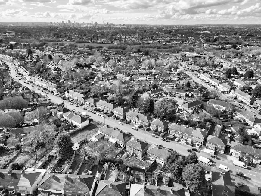 An aerial view of a residential street with a row of terraced houses at the front, each with small front gardens and parked cars along the pavement. Behind the houses, there are multiple fenced garden plots with various features such as lawns, trees, sheds, and patios. Some gardens contain outdoor furniture, play equipment, or garden tools, while others are empty or have construction materials. The scene shows a clear day with natural lighting, highlighting the general environment for house removals or home relocation services. The image illustrates the typical urban setting for furniture transport and packing and moving activities managed by companies like Man with Van North Sheen, emphasizing the importance of proper planning for local property moves and the logistical considerations involved.