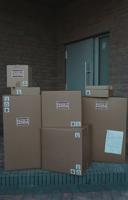 Multiple large cardboard boxes are stacked outside a building beneath a brick wall and near a closed door with glass panels. The boxes vary in size, with some placed directly on a dark brick step and others on a paved surface; they are secured with packing tape and marked with 'fragile' labels and handling symbols. A piece of paper is attached to one of the boxes, possibly an inventory or delivery note. The scene depicts a typical packing stage in the home relocation process, consistent with furniture transport and moving logistics. This setup may be part of the loading process for a house removal service by Man with Van North Sheen. The lighting suggests daytime, and the environment appears prepared for the next steps in the moving operation, highlighting the careful handling of household items during packing and loading.