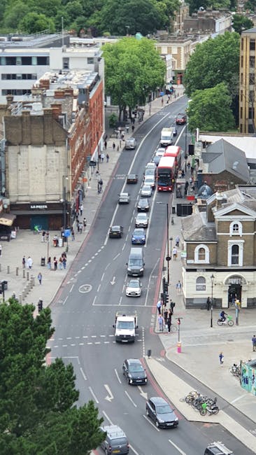 An aerial view of a busy urban street in North Sheen, showing a line of parked vehicles along the curb and a variety of moving cars driving through the area. The street is lined with trees, providing greenery amidst buildings that include residential houses and commercial establishments. Pedestrians are visible on the sidewalks, some walking and others waiting to cross at the pedestrian crossings. The scene highlights the typical environment encountered during home relocation or furniture transport processes, with no movement of large furniture or packing materials visible, but the busy street setting reflects the logistical context of house removals and moving services. Overall, the image captures the lively atmosphere of North Sheen's street scene, relevant for understanding local transport routes involved in professional removals handled by companies like Man with Van North Sheen.